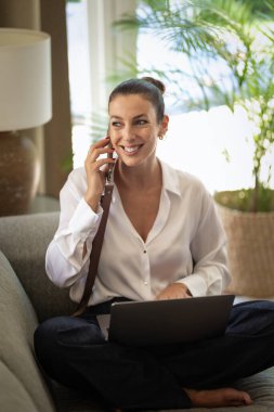 Ofiste dizüstü bilgisayarla çalışırken cep telefonu kullanan gülümseyen bir iş kadını. Esmer, sıradan kıyafetler giyen bir kadın. Tam uzunluk.