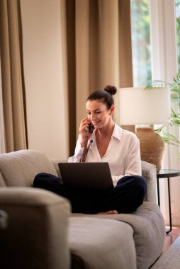 Ofiste dizüstü bilgisayarla çalışırken cep telefonu kullanan gülümseyen bir iş kadını. Esmer, sıradan kıyafetler giyen bir kadın. Tam uzunluk.