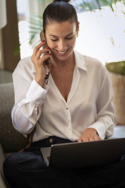 Ofiste dizüstü bilgisayarla çalışırken cep telefonu kullanan gülümseyen bir iş kadını. Esmer kadın, günlük kıyafetler giyiyor.. 