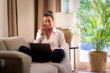 Ofiste dizüstü bilgisayarla çalışırken cep telefonu kullanan gülümseyen bir iş kadını. Esmer, sıradan kıyafetler giyen bir kadın. Tam uzunluk. 