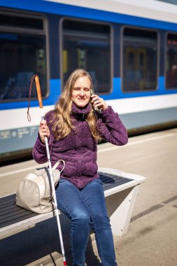 Beyaz bastonlu kör kadın portresi. Şehrin dışındaki tren istasyonunda bankta oturuyor. Treni bekleyen ve telefon kullanan görme engelli bir kadın. Günlük kıyafetler.. 