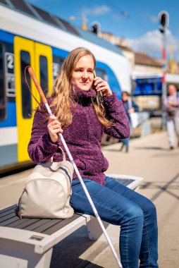Beyaz bastonlu kör kadın portresi. Şehrin dışındaki tren istasyonunda bankta oturuyor. Treni bekleyen ve telefon kullanan görme engelli bir kadın. Günlük kıyafetler.. 