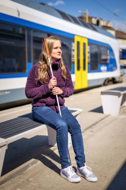 Beyaz bastonlu görme engelli bir kadın tren istasyonunda oturuyor ve birini bekliyor. Tam boy çekim. 
