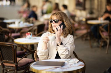 Orta yaşlı bir kadın kaldırımdaki kafede otururken mesaj atıyor. Sarı saçlı, güneş gözlüğü takan ve günlük kıyafetler giyen bir kadın.. 