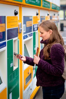 Tren istasyonunda bilet makinesinin önünde durup tren bileti alan görme engelli bir kadın. Kendine güvenen bir kadın işitme cihazı takıyor ve elinde beyaz bir baston tutuyor. Temassız ödeme. 
