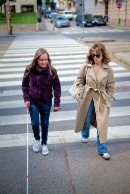 Görme engelli bir kadın yaya geçidinde kadın arkadaşıyla yürüyor. Görme engelli arkadaşına yardım eden olgun, beyaz bir kadın. Tam uzunluk. 