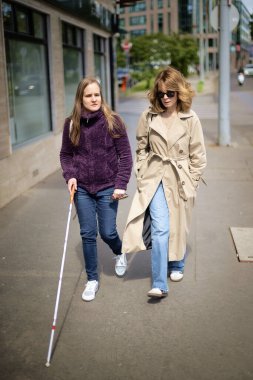 Beyaz bastonlu görme engelli kadın kız arkadaşıyla şehirde yürüyor. Tam boy çekim. 
