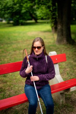 Beyaz baston tutan ve parktaki bankta oturan görme engelli bir kadın. Gülümseyen kadın rahat ceket ve kot pantolon giyiyor.. 