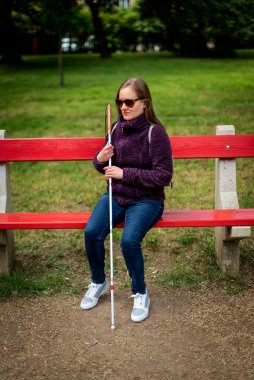 Beyaz baston tutan ve parktaki bankta oturan görme engelli bir kadın. Sıradan ceket ve kot pantolon giyen gülümseyen bir kadın. Tam boy çekim. 