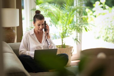 Ofiste dizüstü bilgisayarla çalışırken cep telefonu kullanan gülümseyen bir iş kadını. Esmer, sıradan kıyafetler giyen bir kadın. Tam uzunluk.