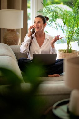 Ofiste dizüstü bilgisayarla çalışırken cep telefonu kullanan gülümseyen bir iş kadını. Esmer, sıradan kıyafetler giyen bir kadın. Tam uzunluk.