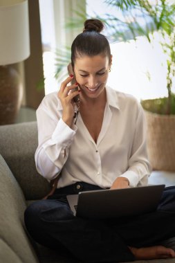 Ofiste dizüstü bilgisayarla çalışırken cep telefonu kullanan gülümseyen bir iş kadını. Esmer, sıradan kıyafetler giyen bir kadın. Tam uzunluk.