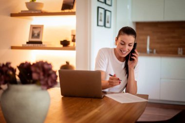 Çekici esmer saçlı kadın modern evinde oturuyor ve çalışıyor. Kendine güvenen bir kadın telefonda konuşuyor ve bir şeyler yazıyor. Sıradan kıyafetler giyen çekici bir iş kadını. Merkez Ofis. 