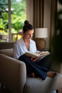 Güzel bir kadın evde rahatlatıcı bir günün tadını çıkarıyor. Orta yaşlı bir kadın rahat koltuğunda oturup kitap okurken gülümsüyor. Tam uzunluk.