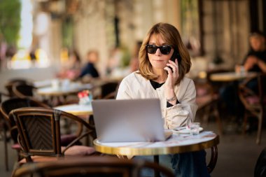 Orta yaşlı bir kadın telefonda biriyle konuşuyor ve kaldırımdaki kafede dizüstü bilgisayar kullanıyor. Sarı saçlı, güneş gözlüğü takan ve günlük kıyafetler giyen bir kadın.. 