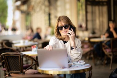 Orta yaşlı bir kadın telefonda biriyle konuşuyor ve kaldırımdaki kafede dizüstü bilgisayar kullanıyor. Sarı saçlı, güneş gözlüğü takan ve günlük kıyafetler giyen bir kadın.. 