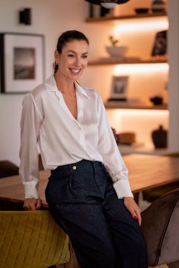 Modern evinde duran esmer saçlı kadın. Neşeli bir gülümseme, kameraya bakmak ve günlük kıyafetler giymek.. 