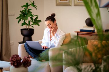 Kendinden emin orta yaşlı bir kadın evden uzakta çalışıyor. Dizüstü bilgisayar kullanırken gülümsüyor. Gündelik görünüş, rahat ve modern iç mekan. Merkez ofis. Serbest çalışma..