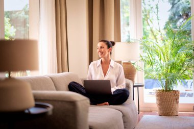 Kendinden emin orta yaşlı bir kadın evden uzakta çalışıyor. Dizüstü bilgisayar kullanırken gülümsüyor. Gündelik görünüş, rahat ve modern iç mekan. Merkez ofis. Serbest çalışma..