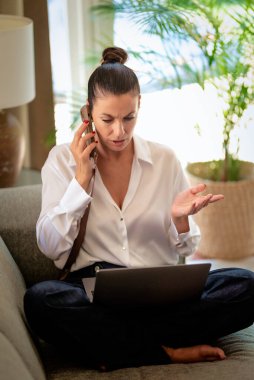 Kendinden emin orta yaşlı bir kadın uzaktan çalışıyor, telefonla konuşuyor ve dizüstü bilgisayar kullanıyor. Sıradan görünüş ve modern iç mekan. Merkez Ofis.