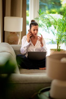 Kendinden emin orta yaşlı bir kadın uzaktan çalışıyor, telefonla konuşuyor ve dizüstü bilgisayar kullanıyor. Sıradan görünüş ve modern iç mekan. Merkez Ofis.