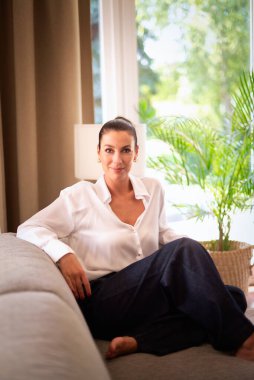 Modern evindeki rahat koltukta oturan orta yaşlı güzel bir kadının tam boy fotoğrafı. Beyaz gömlek ve kot pantolon giyen kendinden emin bir kadın. Tam boy çekim. 