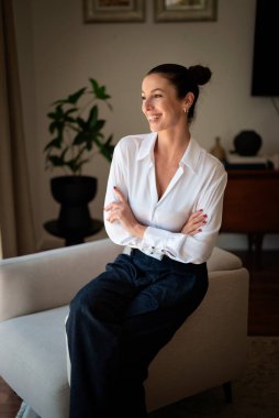 Güzel, orta yaşlı bir kadının koltukta oturup hayal kurduğu bir fotoğraf. Kumral kadın, beyaz gömlek ve kot pantolon giyiyor..