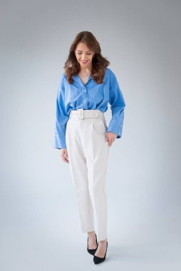 Gülümseyen olgun bir kadının portresi. Açık gri arka planda kumral saçlı, kendinden emin bir kadın. Mavi gömlek ve beyaz pantolon giyen çekici bir kadın. Uzayı kopyala Tam menzil.. 