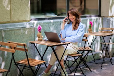Neşeli bir kadın bir masada oturuyor ve açık bir kafede bilgisayar ve akıllı telefon kullanıyor. Sıradan kıyafetler giyen çekici bir kadın. Kendine güvenen bir iş kadını. 