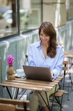 Neşeli kadın bir masada oturuyor ve açık bir kafede bilgisayar kullanıyor. Sıradan kıyafetler giyen çekici bir kadın. Kendine güvenen bir iş kadını. 