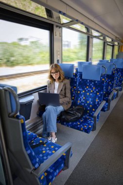 Trenle seyahat ederken dizüstü bilgisayarında çalışan sarışın kadın. Kendine güvenen bir kadın modern bir trende tek başına oturuyor ve dizüstü bilgisayarı üzerinde çalışıyor. Uzaktan kumanda. Tam boy çekim. 