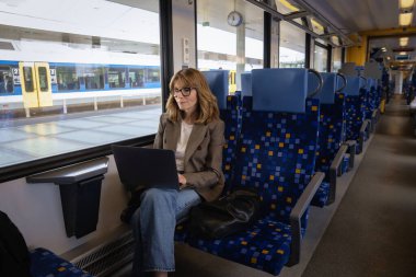 Trenle seyahat ederken dizüstü bilgisayarında çalışan sarışın kadın. Kendine güvenen bir kadın modern bir trende tek başına oturuyor ve dizüstü bilgisayarı üzerinde çalışıyor. Uzaktan kumanda. Tam boy çekim. 