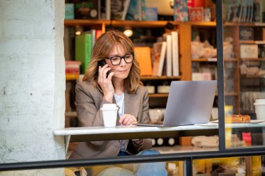 Kendinden emin, orta yaşlı, gözlüklü, stil sahibi bir kadın bir kafedeki masada oturuyor. Yanında duran masada dizüstü bilgisayarı açık ve çalışıyor. Dışarıdan çekilmiş bir fotoğraf. Uzak çalışma. 