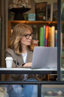Kendinden emin, orta yaşlı, gözlüklü, stil sahibi bir kadın bir kafedeki masada oturuyor. Yanında duran masada dizüstü bilgisayarı açık ve çalışıyor. Dışarıdan çekilmiş bir fotoğraf. Uzak çalışma. 