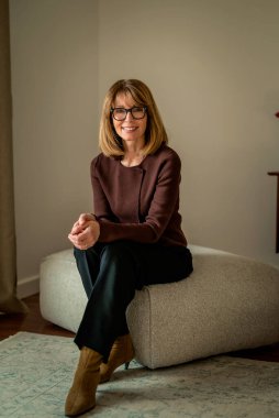 Duvarın önünde oturan gülümseyen kadın. Sıradan kıyafetler ve gözlük takan çekici bir kadın. Sarı saçlı kadın kameraya gülümsüyor. Uzayı kopyala Tam uzunluk.