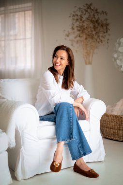 Güzel bir kadın evde rahatlatıcı bir günün tadını çıkarıyor. Orta yaşlı kadın rahat koltuğunda otururken ışıl ışıl gülümsüyor. Tam boy çekim. 