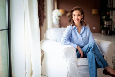 Güzel bir kadın evde rahatlatıcı bir günün tadını çıkarıyor. Orta yaşlı kadın rahat koltuğunda otururken ışıl ışıl gülümsüyor. Tam boy çekim. 