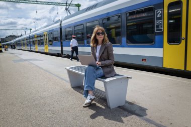 Tren istasyonunda elinde dizüstü bilgisayarla oturan orta yaşlı bir kadın. İş kıyafetleri ve güneş gözlüğü takan kendinden emin bir kadın. Tam uzunluk. 