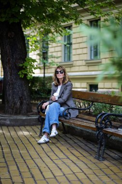 Güneşli bir günde orta yaşlı bir kadın bankta oturuyor. Kendine güvenen kadın spor ceket giyiyor ve gözlüklü kot pantolon giyiyor. Elinde kağıt kahve fincanı vardı. Kahve molası. Tam uzunluk. 