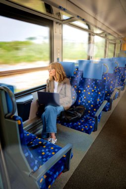 Dizüstü bilgisayarı olan sarı saçlı kadın trenle seyahat ediyor ve pencereden dikkatlice bakıyor. Kendine güvenen bir kadın modern bir trende tek başına oturuyor ve dizüstü bilgisayarı üzerinde çalışıyor. Uzaktan kumanda. Tam boy çekim. 