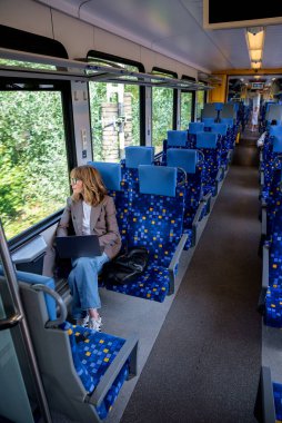 Dizüstü bilgisayarı olan sarı saçlı kadın trenle seyahat ediyor ve pencereden dikkatlice bakıyor. Kendine güvenen bir kadın modern bir trende tek başına oturuyor ve dizüstü bilgisayarı üzerinde çalışıyor. Uzaktan kumanda. Tam boy çekim. 