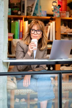 Kendinden emin, orta yaşlı, gözlüklü, stil sahibi bir kadın bir kafedeki masada oturuyor. Yanında duran masada dizüstü bilgisayarı açık ve çalışıyor. Dışarıdan çekilmiş bir fotoğraf. Uzak çalışma. 