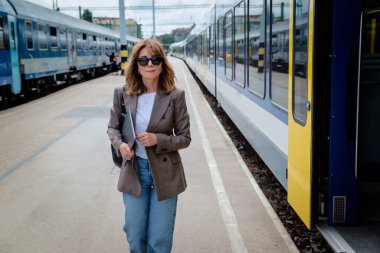 İki tren arasındaki tren peronunda elinde defterle yürüyen kendinden emin orta yaşlı bir kadın. Sarı saçlı, şık giyinen ve kameraya bakan bir kadın. Banliyö kadını işe gidiyor..