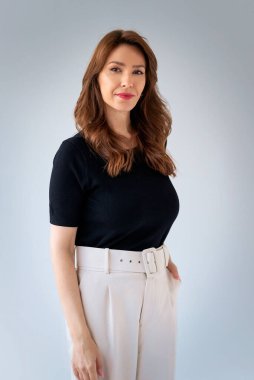 Gülümseyen olgun bir kadının portresi. Beyaz arka planda esmer saçlı, kendinden emin bir kadın. Siyah kazak ve beyaz pantolon giyen çekici bir kadın. Boşluğu kopyala. 