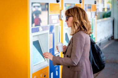 Tren istasyonunda bilet makinesinin önünde duran kendinden emin, orta yaşlı bir kadın. Kendine güveni tam bir kadın iş kıyafeti giyiyor ve elinde kağıt bir kahve fincanı tutuyor.. 