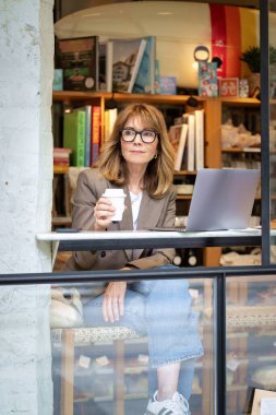 Kendinden emin, orta yaşlı, gözlüklü, stil sahibi bir kadın bir kafedeki masada oturuyor. Yanında duran masada dizüstü bilgisayarı açık ve çalışıyor. Dışarıdan çekilmiş bir fotoğraf. Uzak çalışma. 