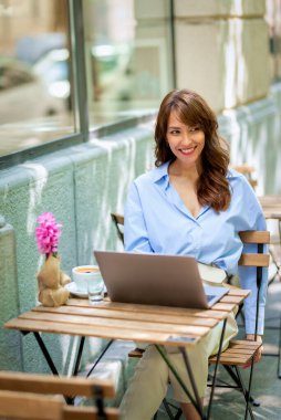 Neşeli kadın bir masada oturuyor ve açık bir kafede bilgisayar kullanıyor. Sıradan kıyafetler giyen çekici bir kadın. Kendine güvenen bir iş kadını çalışıyor. Uzak çalışma. 