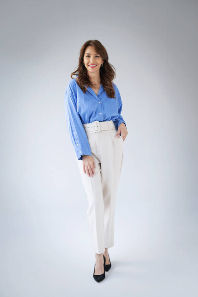 Портрет улыбающейся зрелой женщины. Уверенная женщина с брюнетками на белом фоне. Привлекательная женщина в голубой рубашке и белых штанах. Принято. Полная длина. 