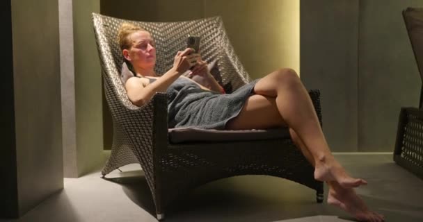 穿着毛巾的年轻女人晚上在游泳池边放松地躺在椅子上 女孩用智能手机给她的朋友打字 高质量的4K镜头 — 图库视频影像
