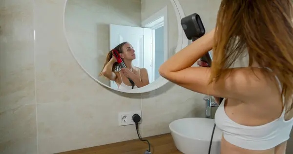 Woman looking at her reflection in the mirror and drying hair with styler after taking shower in bathroom. High quality photo